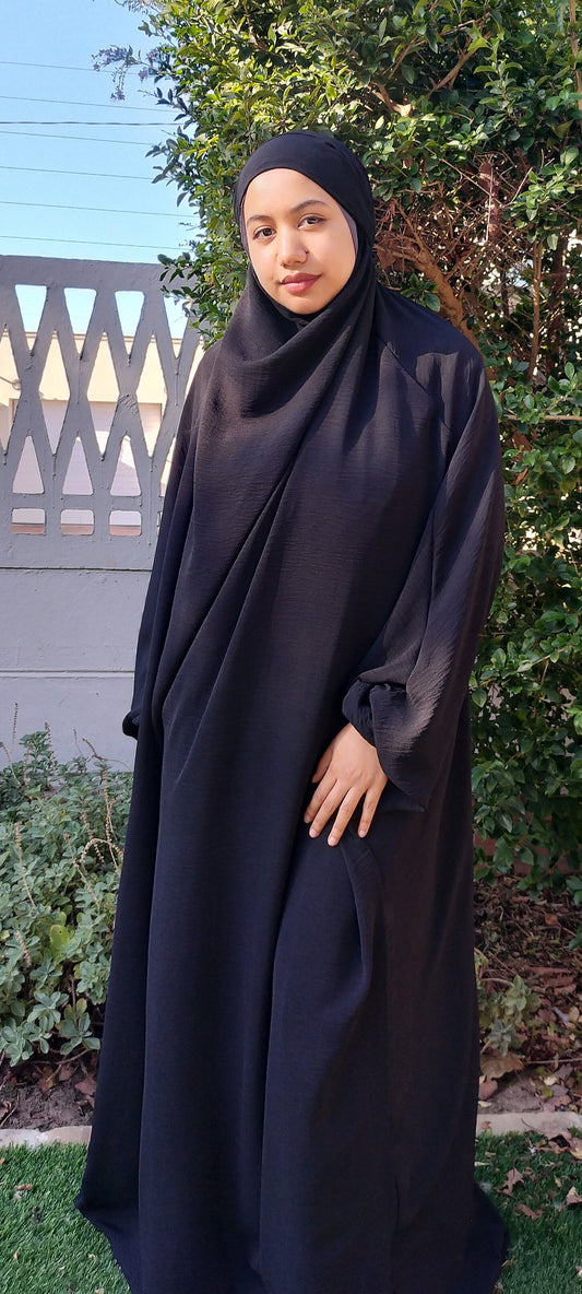 Person wearing a black abaya standing outdoors with greenery in the background