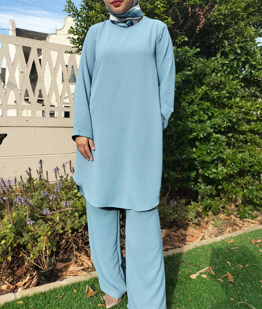 Person wearing a light blue outfit standing outdoors with greenery in the background