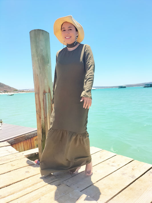 Woman in a long green dress and hat standing on a wooden dock by a turquoise body of water.