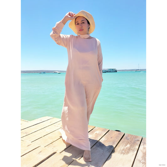 Woman in a long nude modest dress and hat standing on a wooden dock by a body of water. Modelling the Regal Couture modest fashion collection.